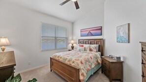 Ein Schlafzimmer mit einem hölzernen Bett, einem Nachttisch, einer Lampe, einem Fenster mit Jalousien, einem Deckenventilator und Kunstwerken an den Wänden.