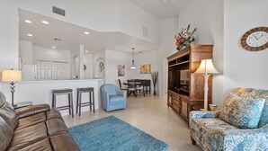 A spacious living room with a brown leather sofa, a blue armchair, a patterned sofa, a dining area with a table and chairs, a TV stand, and a kitchen area in the background. - Lakeview Lanai in Fort Myers! Gated community with resort like amenities!!! (Fort Myers)