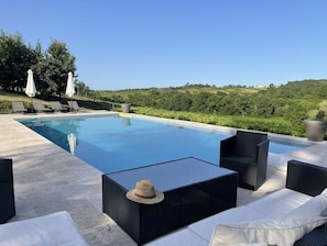 Una piscina con sillones de descanso y una mesa, rodeada de vegetación y colinas.