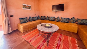 A living room with a sofa set, a round table with a bowl of fruit, and a flat-screen TV mounted on the wall.