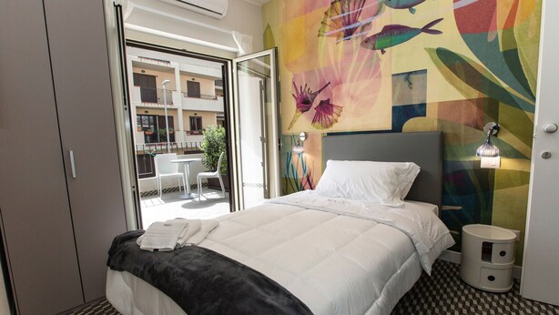 A hotel room with a bed, a white pillow, a black throw blanket, a bedside table with a lamp, and a colorful mural on the wall. - Malu Suites (Messina)