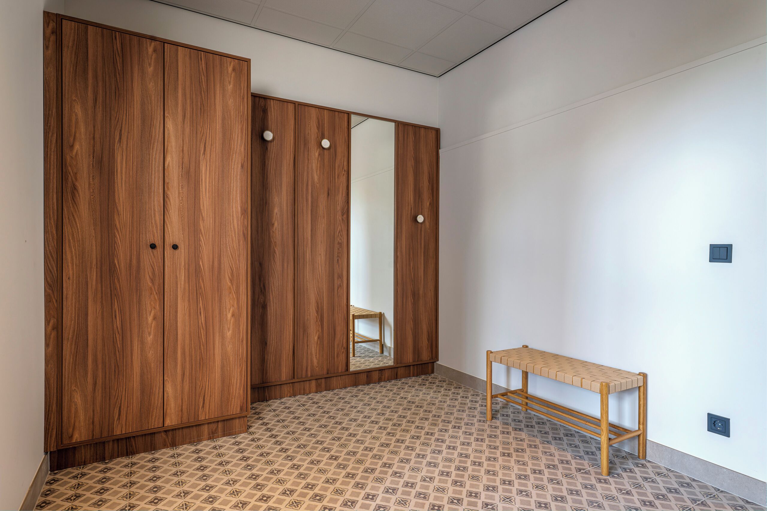A room with a large wooden wardrobe, a small wooden bench, and patterned floor tiles.