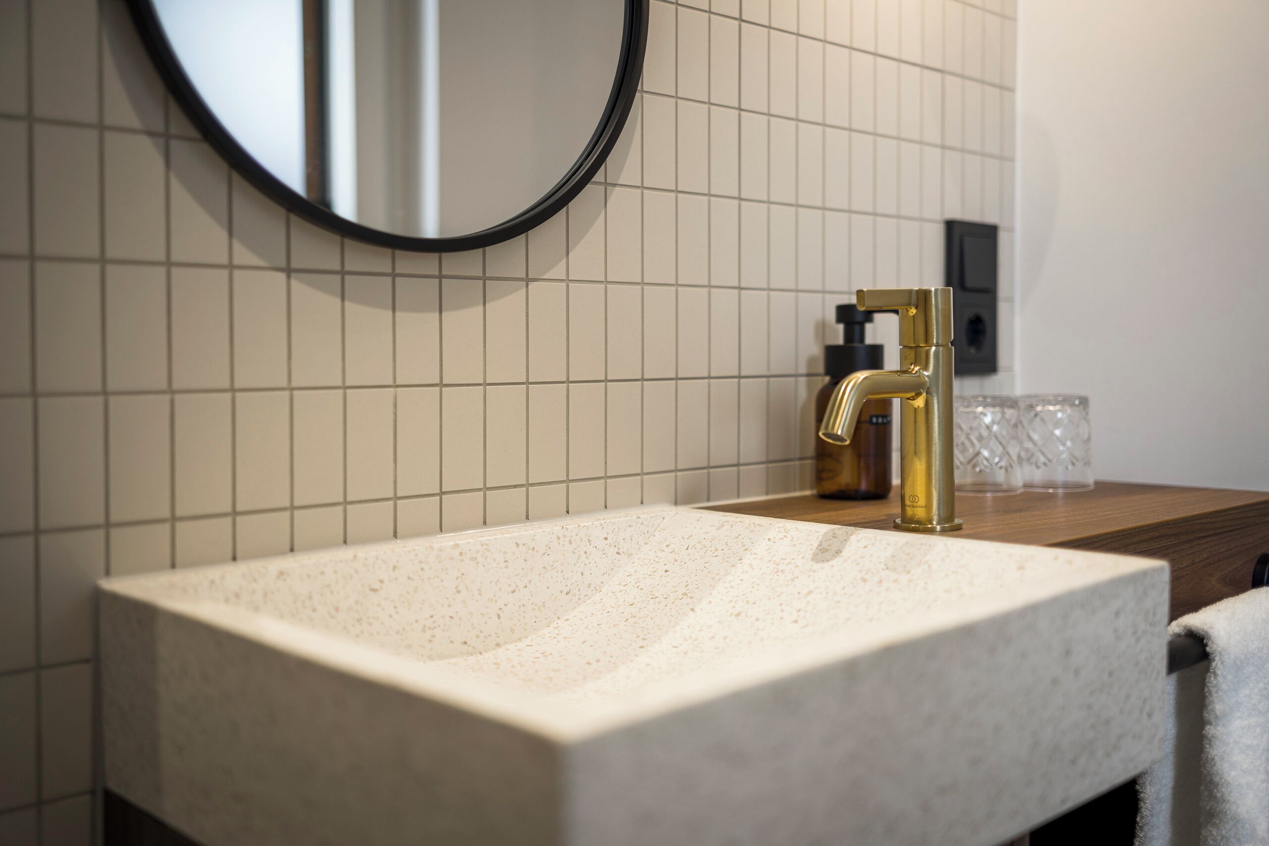 A bathroom with a white speckled sink, a gold faucet, a round mirror, and a tiled wall.
