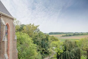 Edifício de tijolos com uma grande janela em arco e vista para uma paisagem verde com árvores e campos.