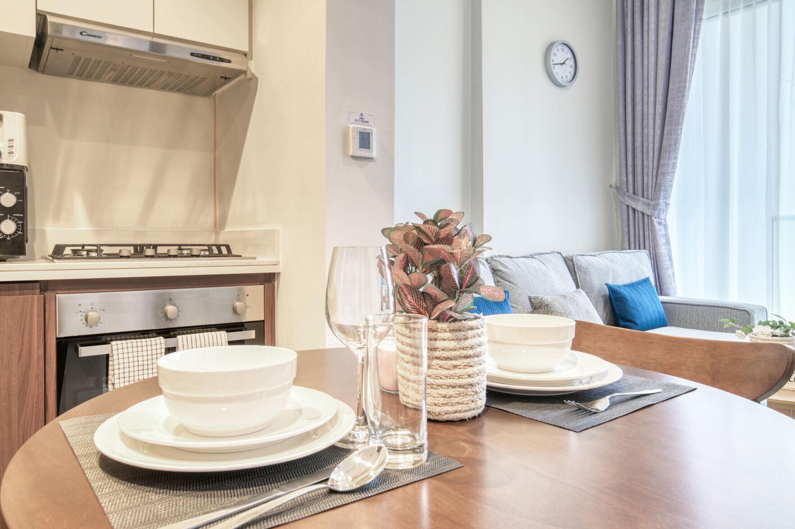 Un comedor con una mesa redonda dispuesta para dos personas, una cocina con estufa a gas, y una sala de estar con un sofá y un reloj en la pared.