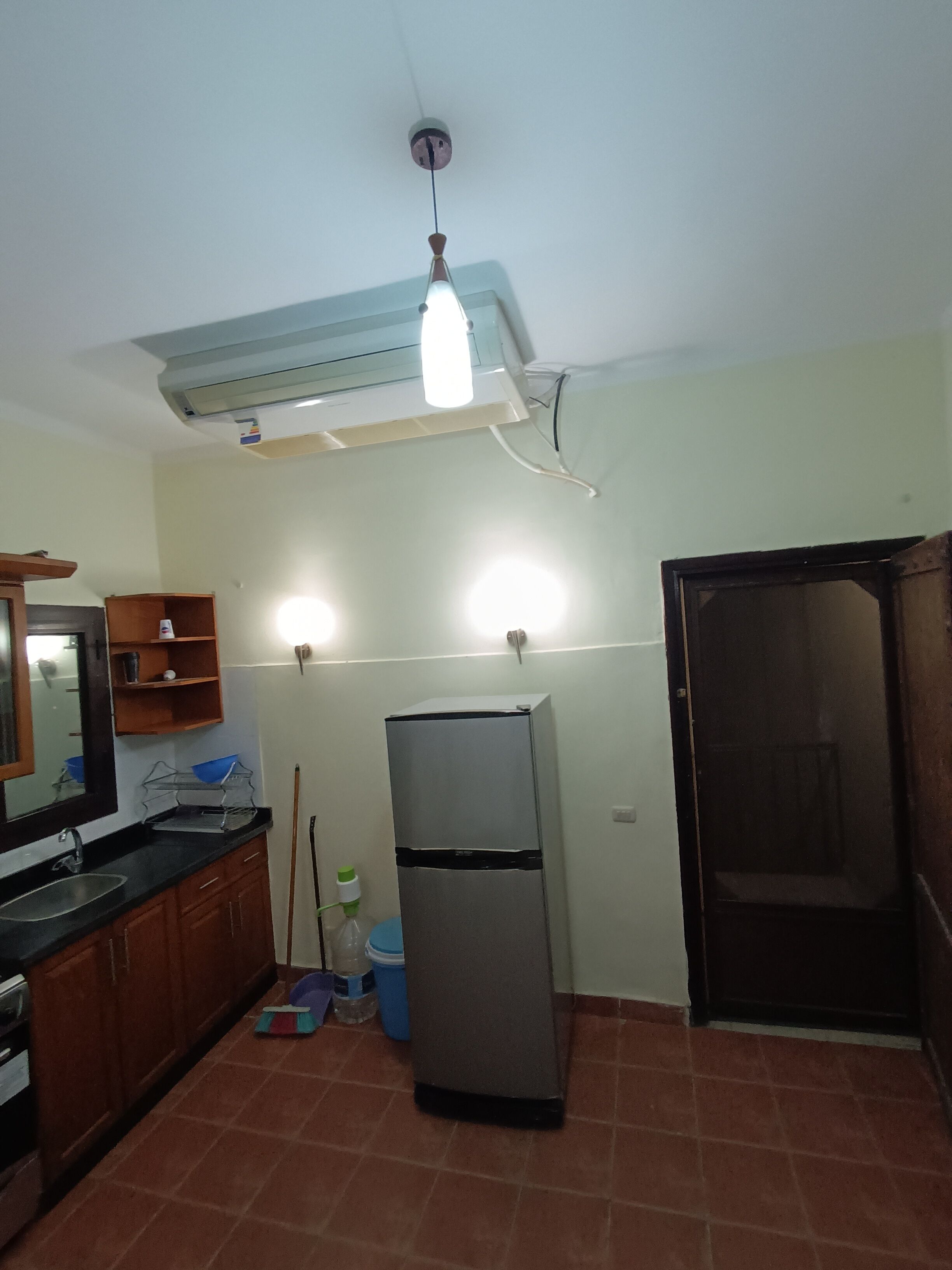Une cuisine de taille réduite équipée d’un réfrigérateur, d’un évier et d’une cuisinière. Un ventilateur de plafond et un luminaire sont installés au-dessus du plan de travail.