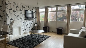A living room with a fireplace, a TV mounted on the wall, a sofa, a dining table, and a view of trees through large windows.