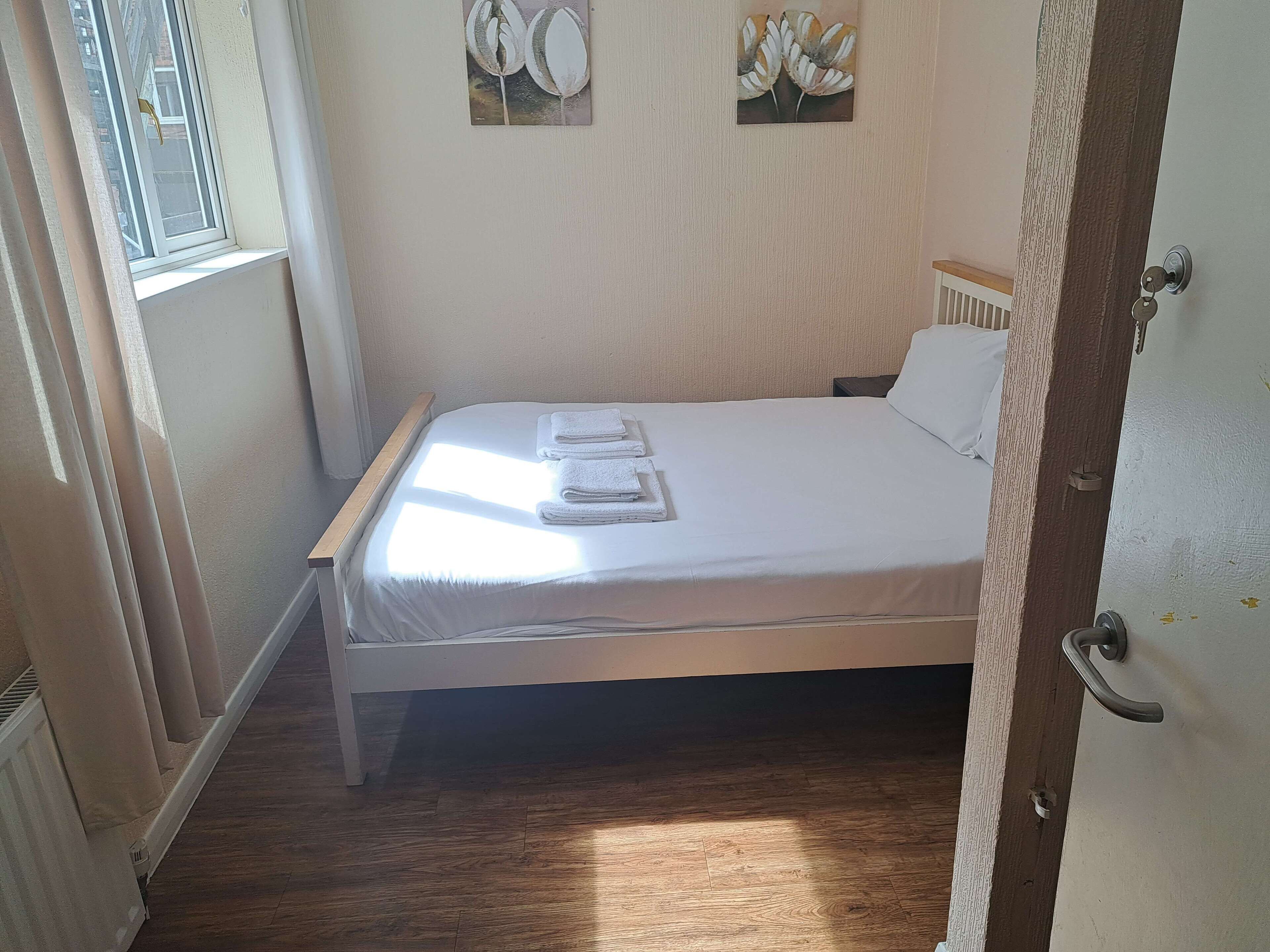 A neatly made bed with white linens and towels, a window with curtains, and two wall-mounted paintings.
