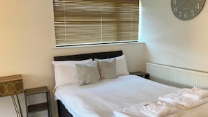 A neatly made bed with white linens, a bedside table with a lamp, and a wall clock.