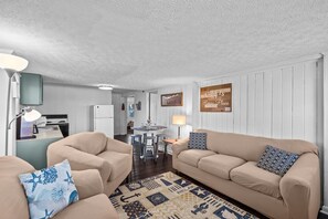 Una sala acogedora con dos sofás beige, un tapete estampado y una cocina al fondo.