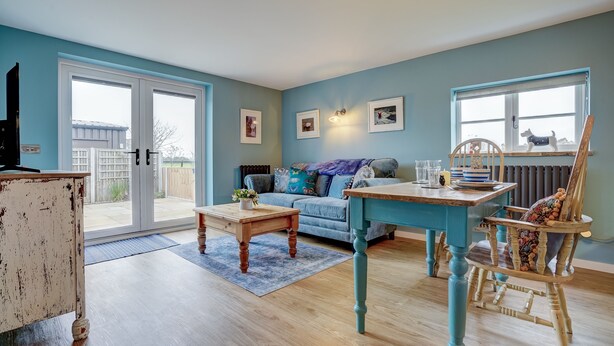 A living room with a blue sofa, a wooden coffee table, a dining table with chairs, and a large window with a view of the outside. - The Peanut | East Ruston Cottages (Happisburgh)