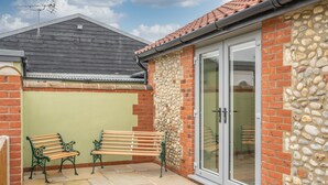 A patio with two wooden benches, a stone wall, and a glass door leading to an interior space. - Bumblebee Cottage | East Ruston Cottages (Happisburgh)