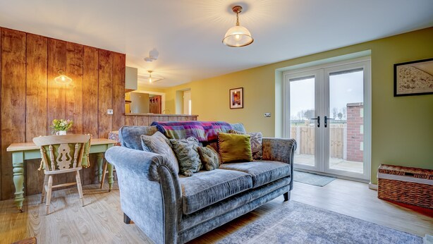 A cozy living room with a grey sofa, wooden wall paneling, and a dining area with a green table and a chair. - Bumblebee Cottage | East Ruston Cottages (Happisburgh)