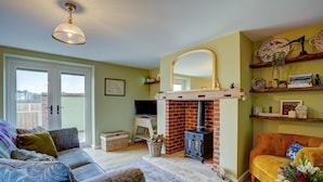 A cozy living room with a fireplace, a sofa, a yellow armchair, and a television. - Bumblebee Cottage | East Ruston Cottages (Happisburgh)