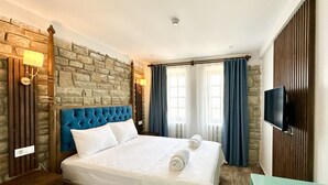 Una habitación de hotel con pared de piedra, cabecero azul acolchado, cama con ropa blanca, televisor montado en la pared y mesita de noche de madera.