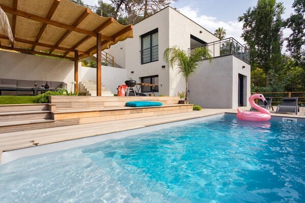 Un área de piscina moderna con cubierta de madera, un flotador de flamenco rosa y una zona de estar techada.
