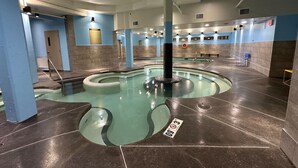 Een groot binnenzwembad met een centrale paal, omringd door een betegelde vloer en muren in blauw en beige tinten.