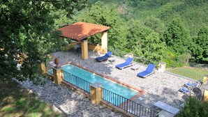 A swimming pool with lounge chairs and a covered seating area, surrounded by greenery and stone flooring.