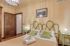 A bedroom with a wooden door, a bed with green and white bedding, two bedside tables with lamps, a chandelier, and a framed picture on the wall.