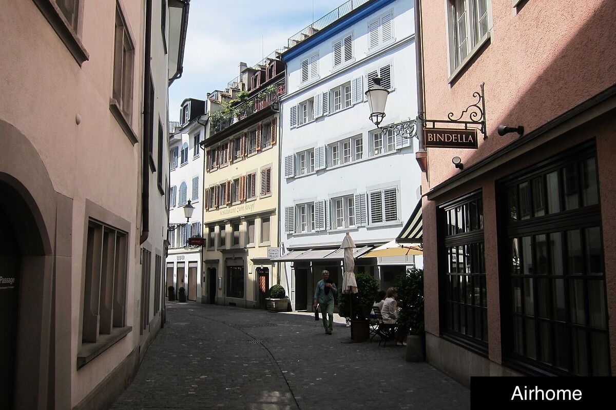 Eine schmale Straße mit Häusern an beiden Seiten, ein Schild mit 'BINDELLA' über einem Geschäft, und eine Person, die geht.