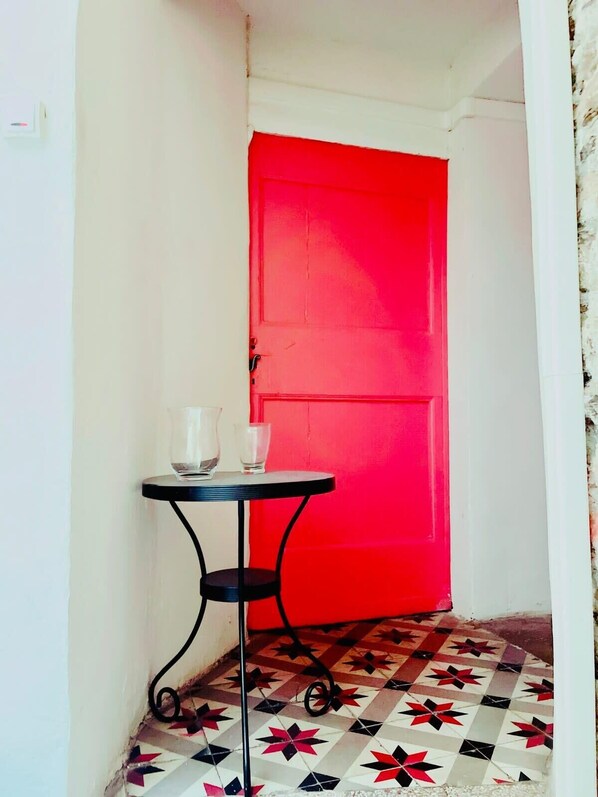 A narrow hallway with a red door, a small table with two glasses, and patterned floor tiles. - Résidence St Vincent (Collioure)
