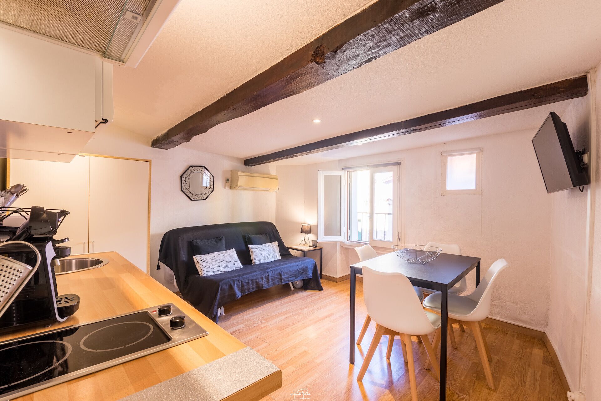 A compact living space with a kitchenette, a sofa, a dining table, and a TV mounted on the wall.