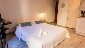 A neatly made bed with white bedding and two black and white pillows, a small bedside table with a lamp, and a wooden floor. - Résidence St Vincent (Collioure)