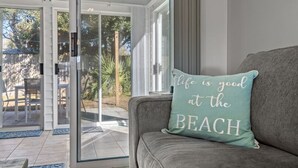 Un salón con un sofá gris, un cojín verde azulado con la frase "Life is good at the BEACH", y un ventanal de vidrio que da a un patio exterior con una mesa y sillas.