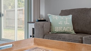 Una sala de estar con un sofá gris, un cojín con la inscripción "life is good at the beach" y una ventana que ofrece vistas a una cerca y follaje.