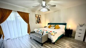 Un dormitorio con cama, un banco, un ventilador de techo, un aparador y una ventana con cortinas.
