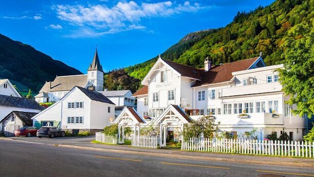 A row of white buildings with a church steeple, a white picket fence, and a mountain backdrop. - Utne Hotel (Ullensvang)