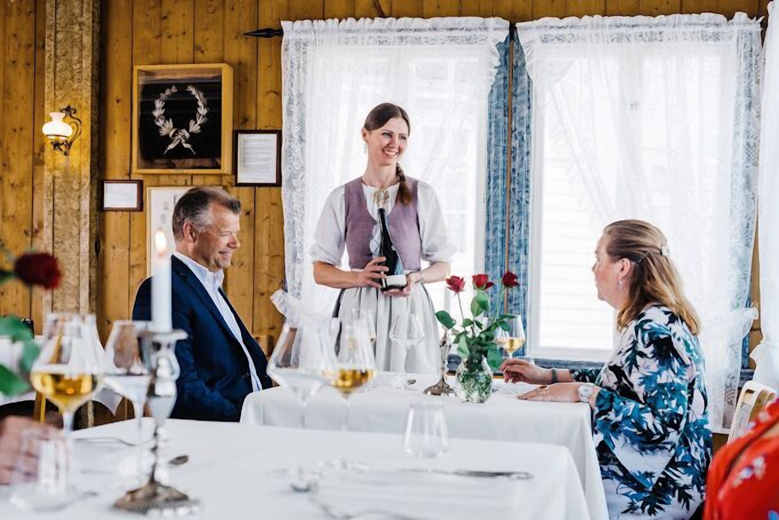 A wooden interior with a dining table set for a meal, a waiter, and guests.