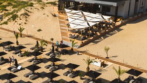 Vista aérea de un complejo turístico frente a la playa, con numerosas tumbonas y sombrillas, una zona de estar cubierta y un edificio con tejado plano.