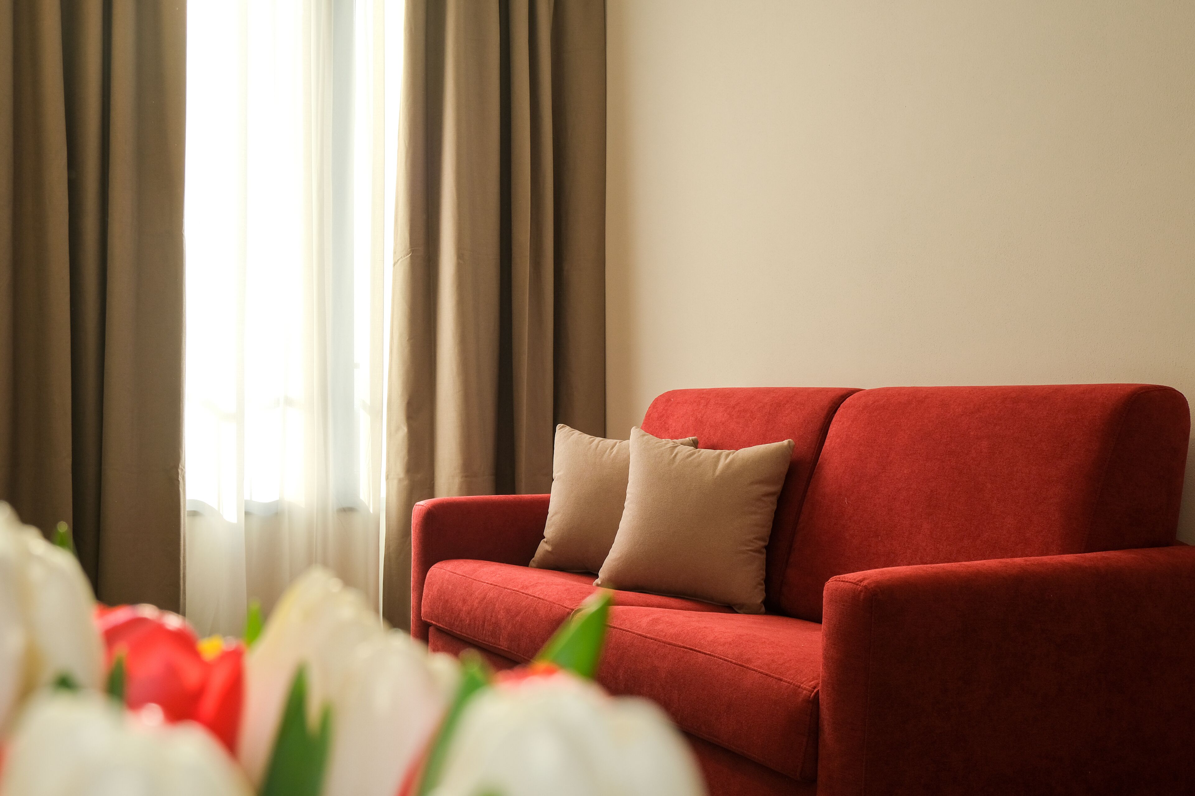 A red sofa with beige cushions in a room with a window and curtains.