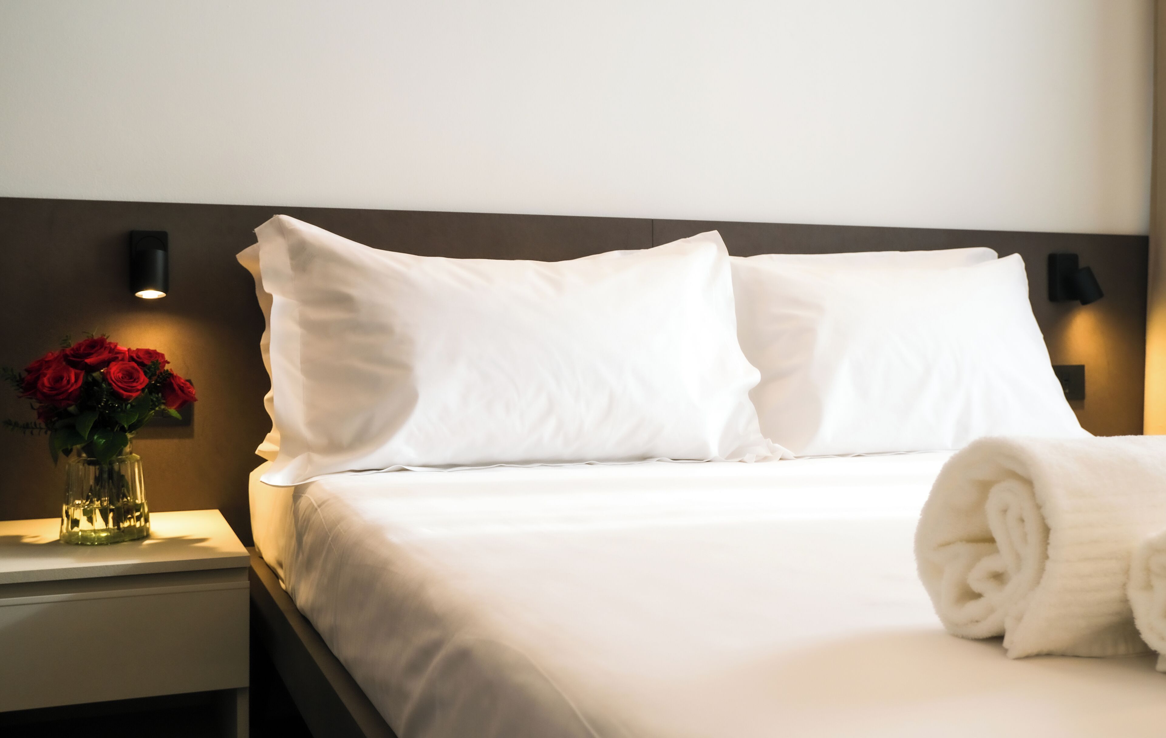 A neatly made bed with white linens, a vase of red roses on the nightstand, and a wall-mounted light fixture.