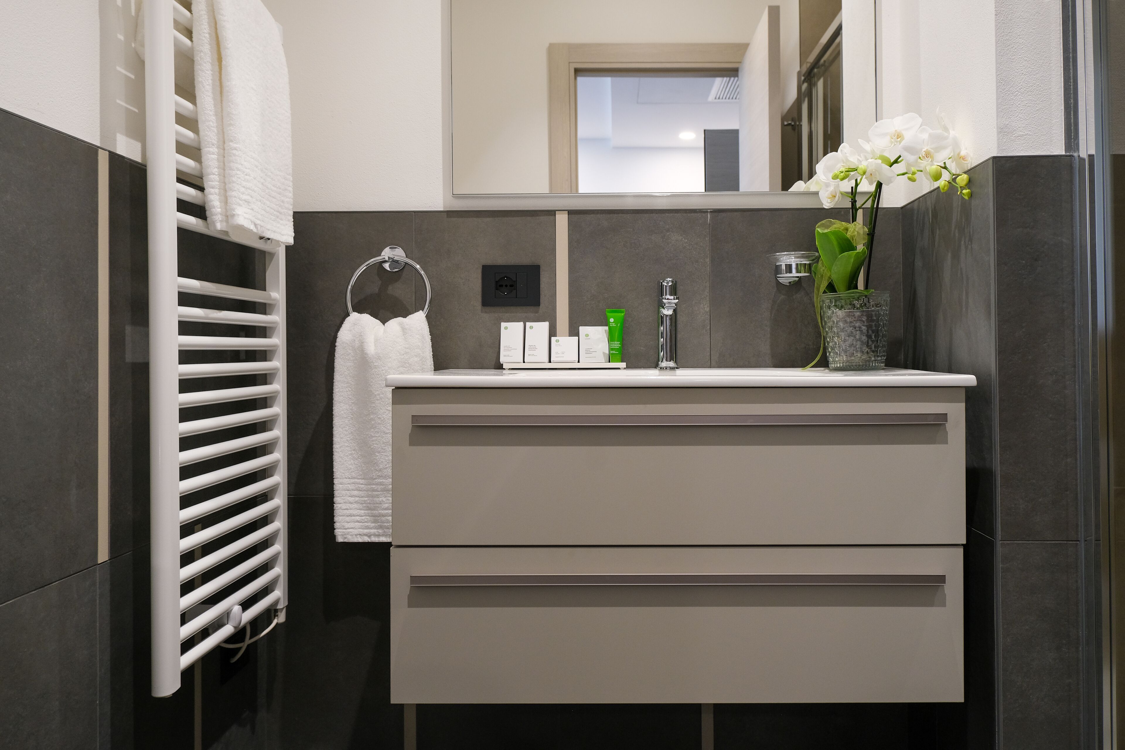 A modern bathroom with a white vanity, a towel rack, a mirror, and a vase with flowers.