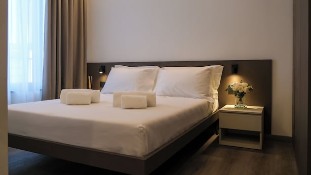 A neatly made bed with white linens and two towels, a bedside table with a vase of flowers, and a wall-mounted lamp. - Emerald Green Residence (Caldiero)