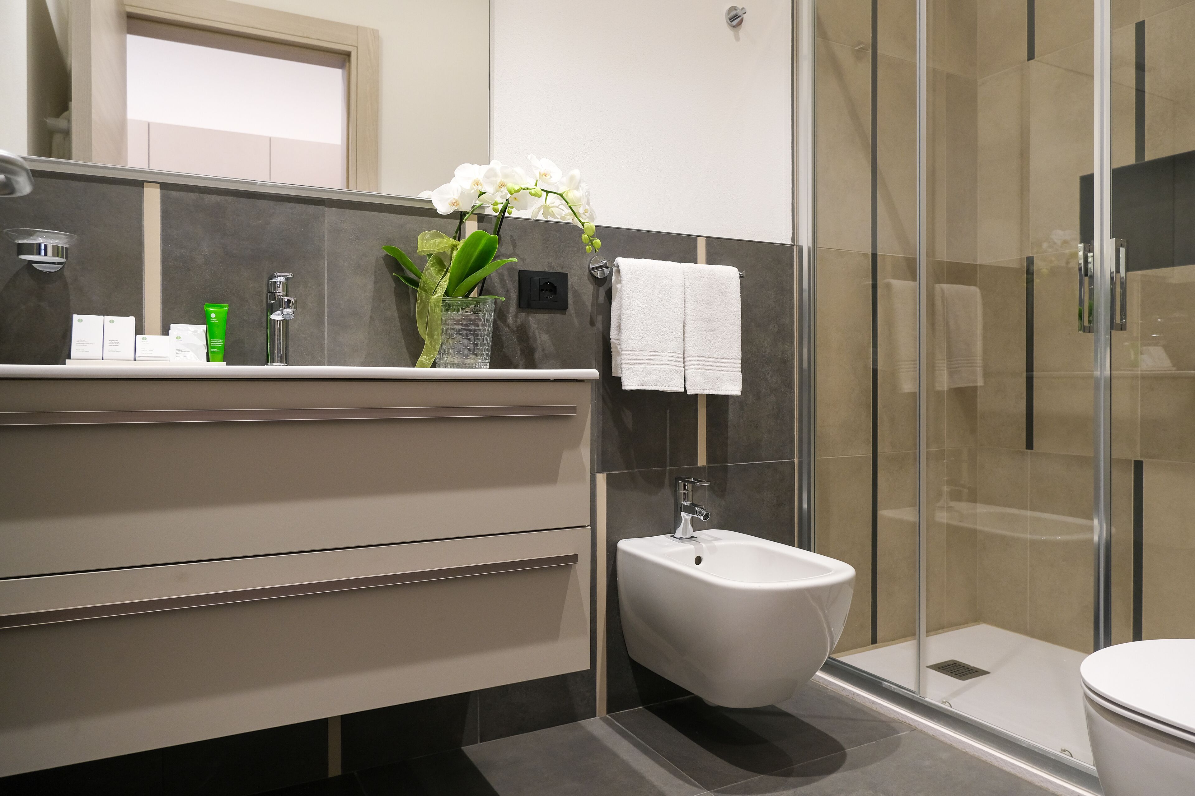 Ein modernes Badezimmer mit integriertem Bidet, einer Duschkabine aus Glas, einem Waschtisch mit Spiegel und einem Topf mit einer Orchidee.