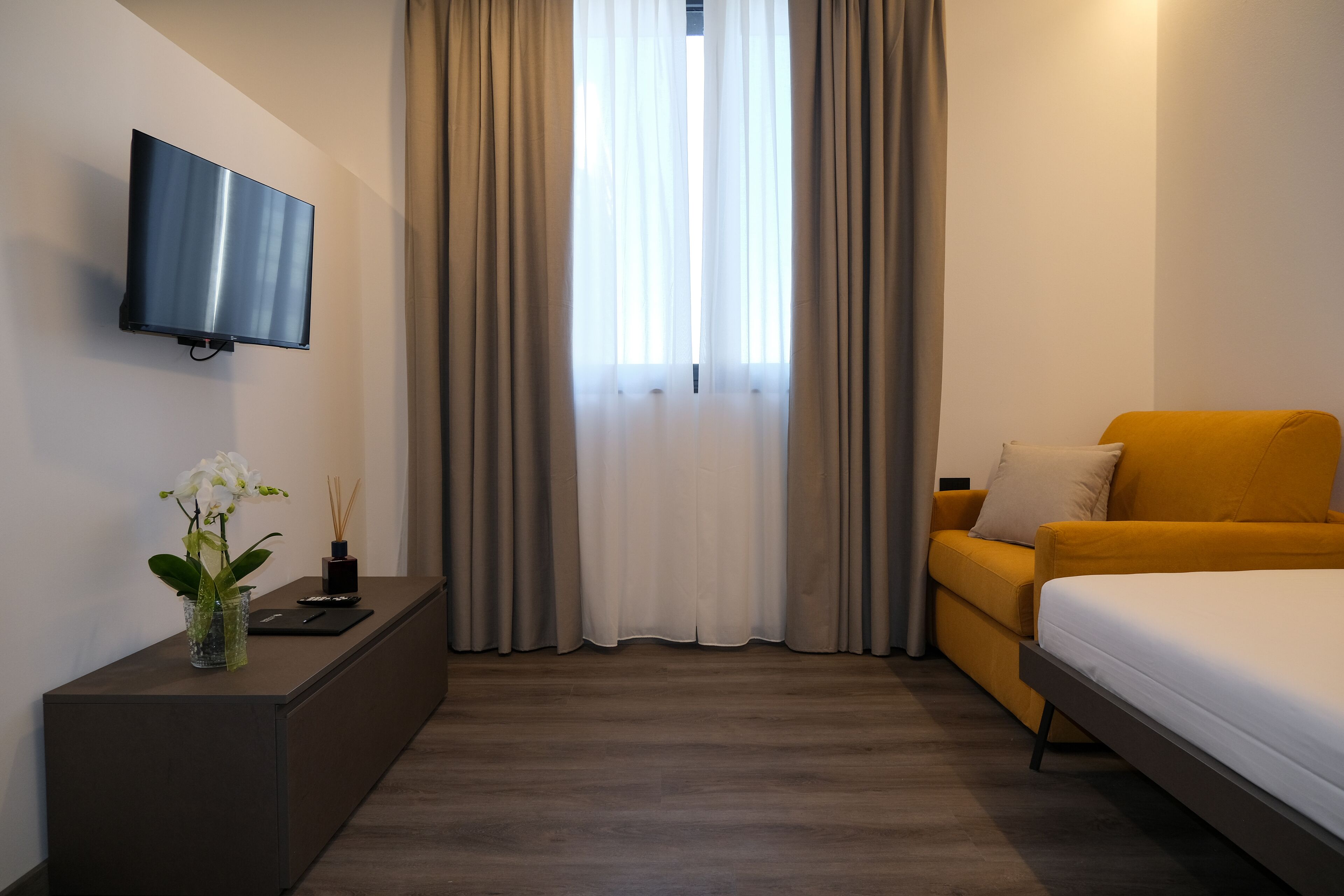 A hotel room with a flat-screen TV mounted on the wall, a sofa, a bed, a small table with a vase of flowers, and a window with curtains.