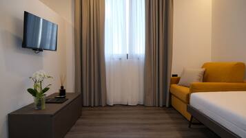 A hotel room with a flat-screen TV mounted on the wall, a sofa, a bed, a small table with a vase of flowers, and a window with curtains.
