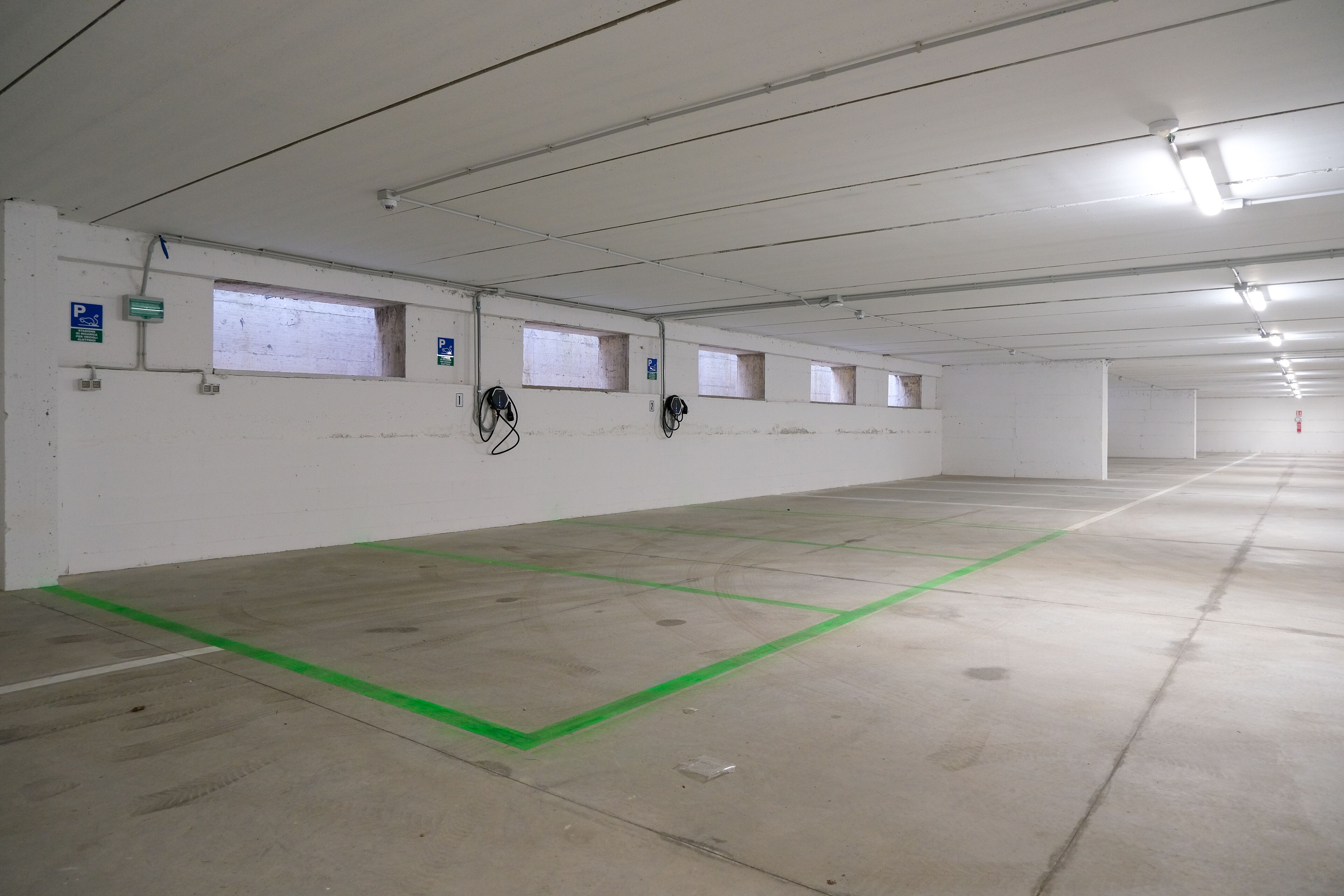 Un grand parking souterrain, vide, avec un sol en béton, des lignes vertes délimitant les places et de nombreuses fenêtres le long des murs.