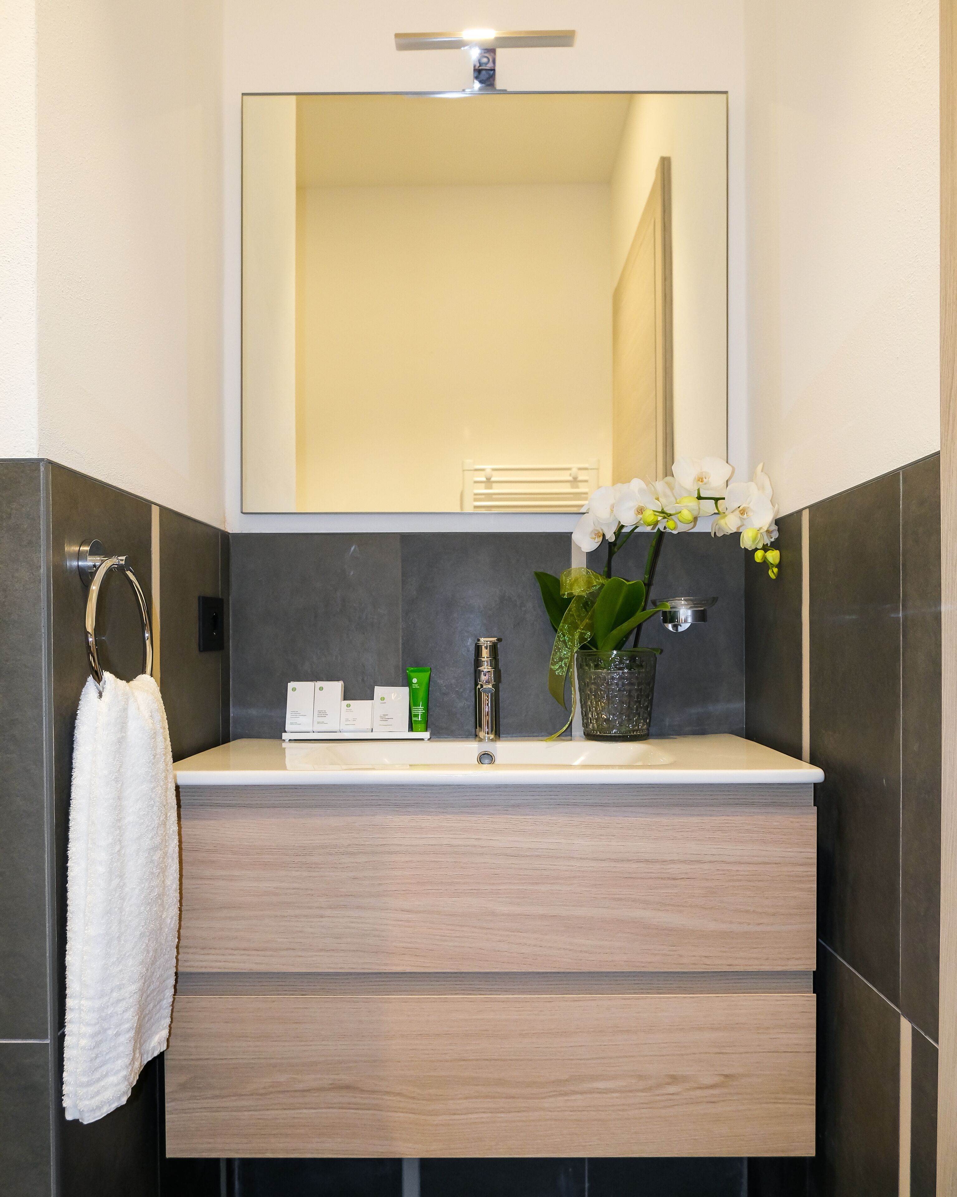 Un baño moderno con un mueble de tocador de madera, un espejo, un perchero para toallas y una orquídea en maceta.