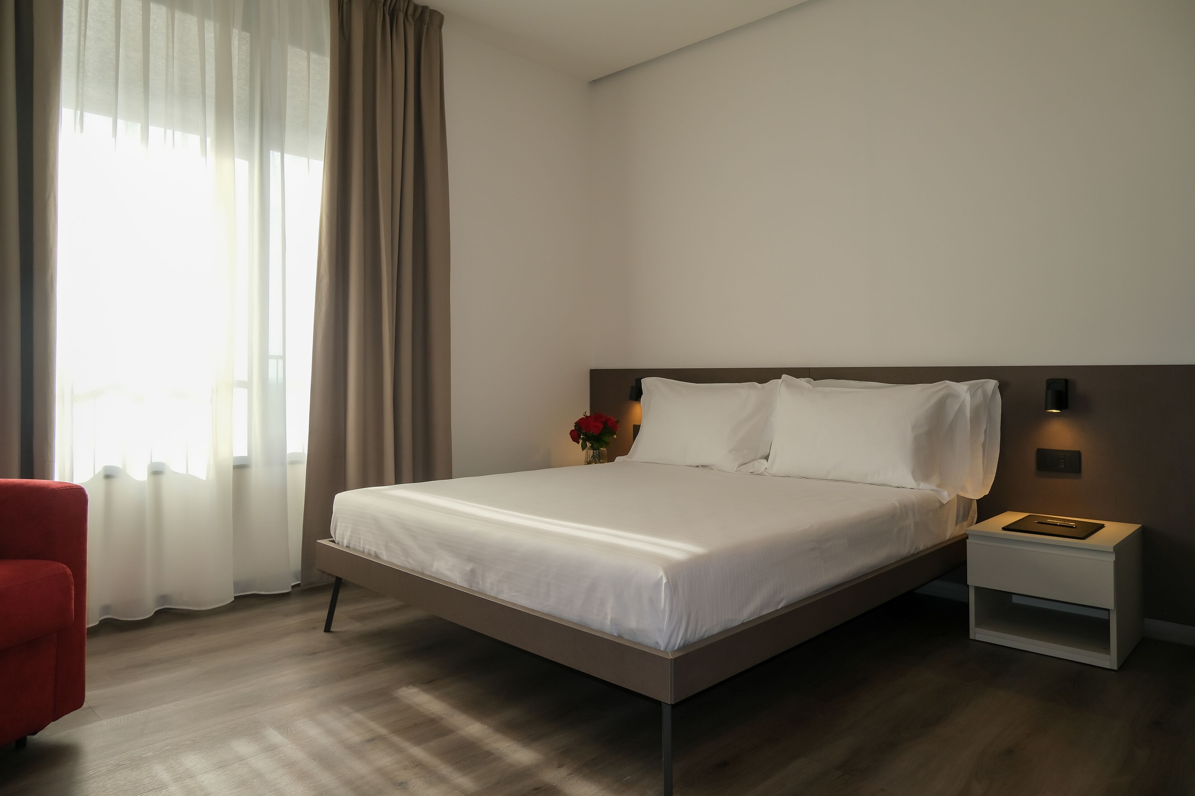 A modern hotel room with a large bed, a bedside table with a lamp, a red chair, and a window with sheer curtains.