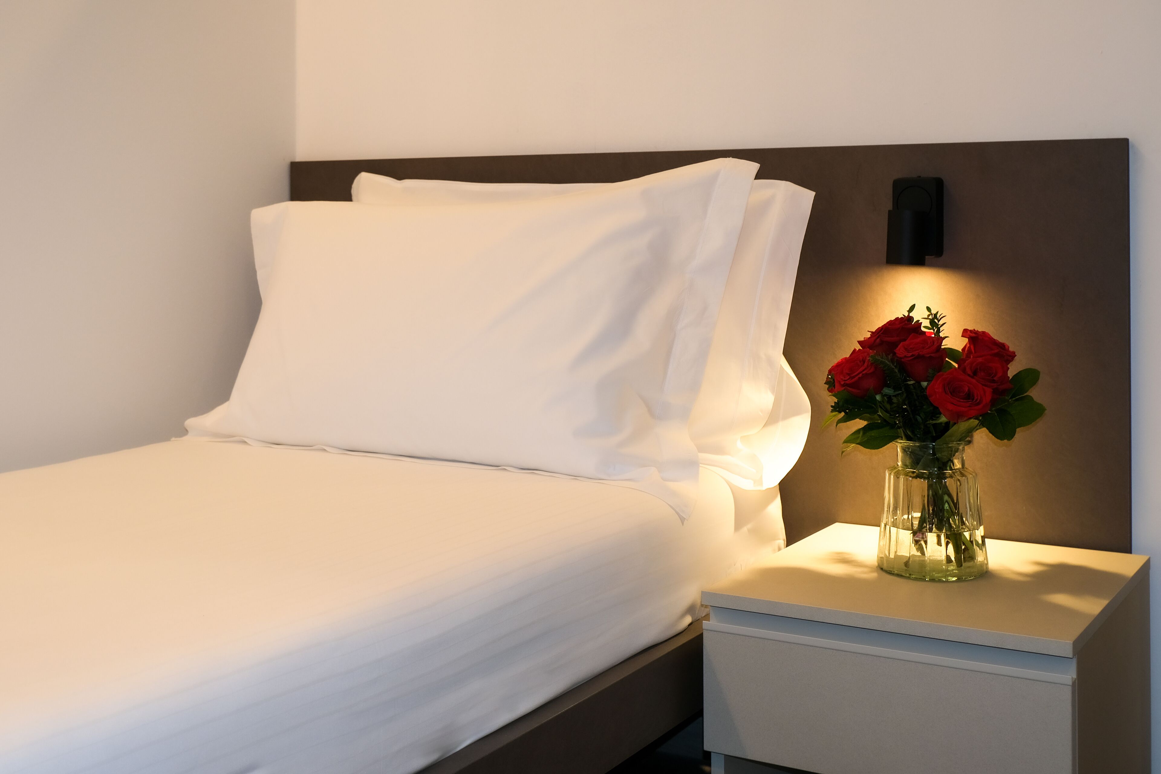 A neatly made bed with white linens, a bedside table with a vase of red roses, and a wall-mounted light fixture.