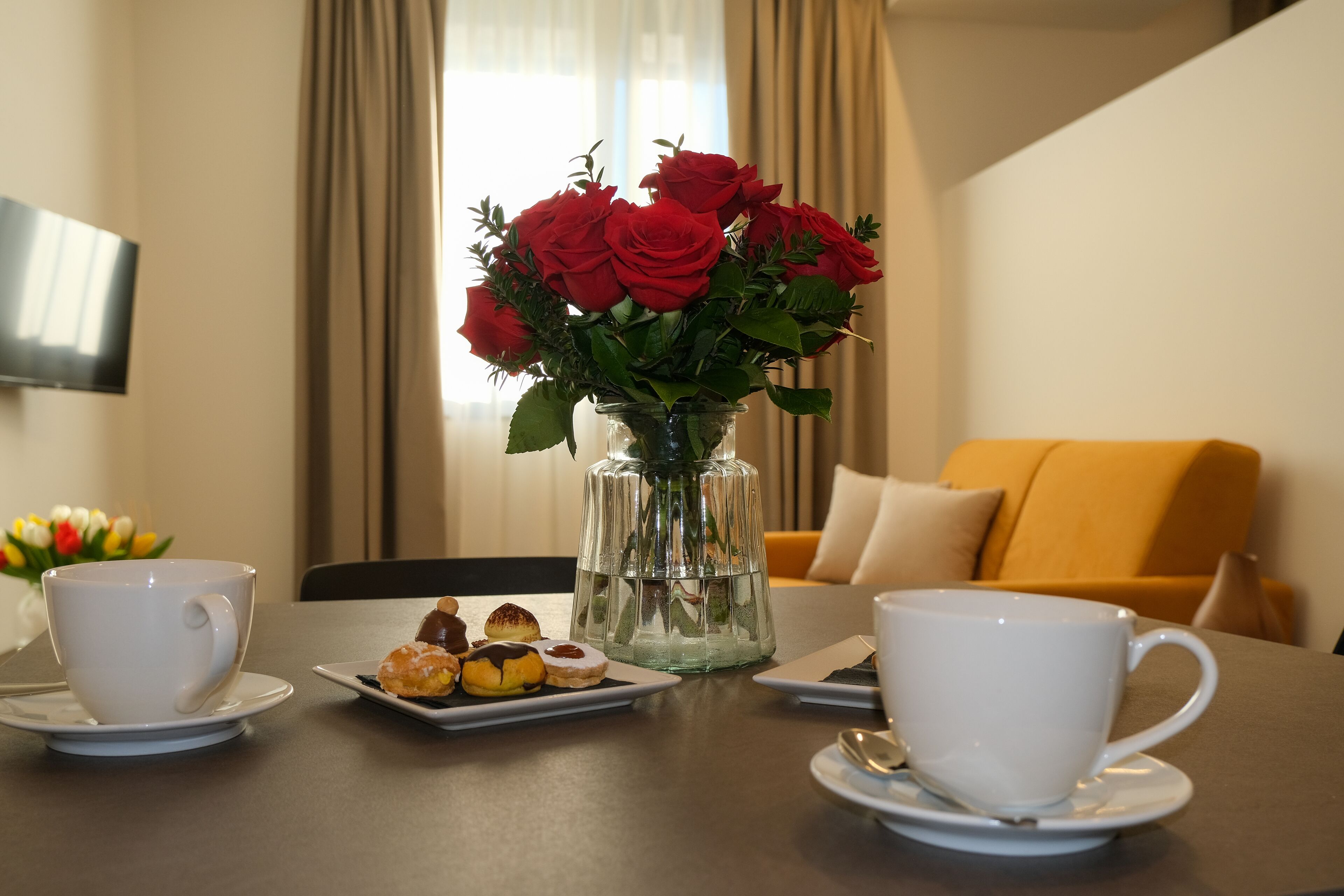 A dining table with two white cups, a glass vase with red roses, and a plate of pastries.
