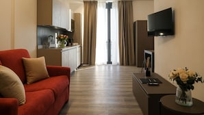 A modern hotel room with a red sofa, a small table with a vase of flowers, a flat-screen TV mounted on the wall, and a kitchen area visible through a glass door. - Emerald Green Residence (Caldiero)