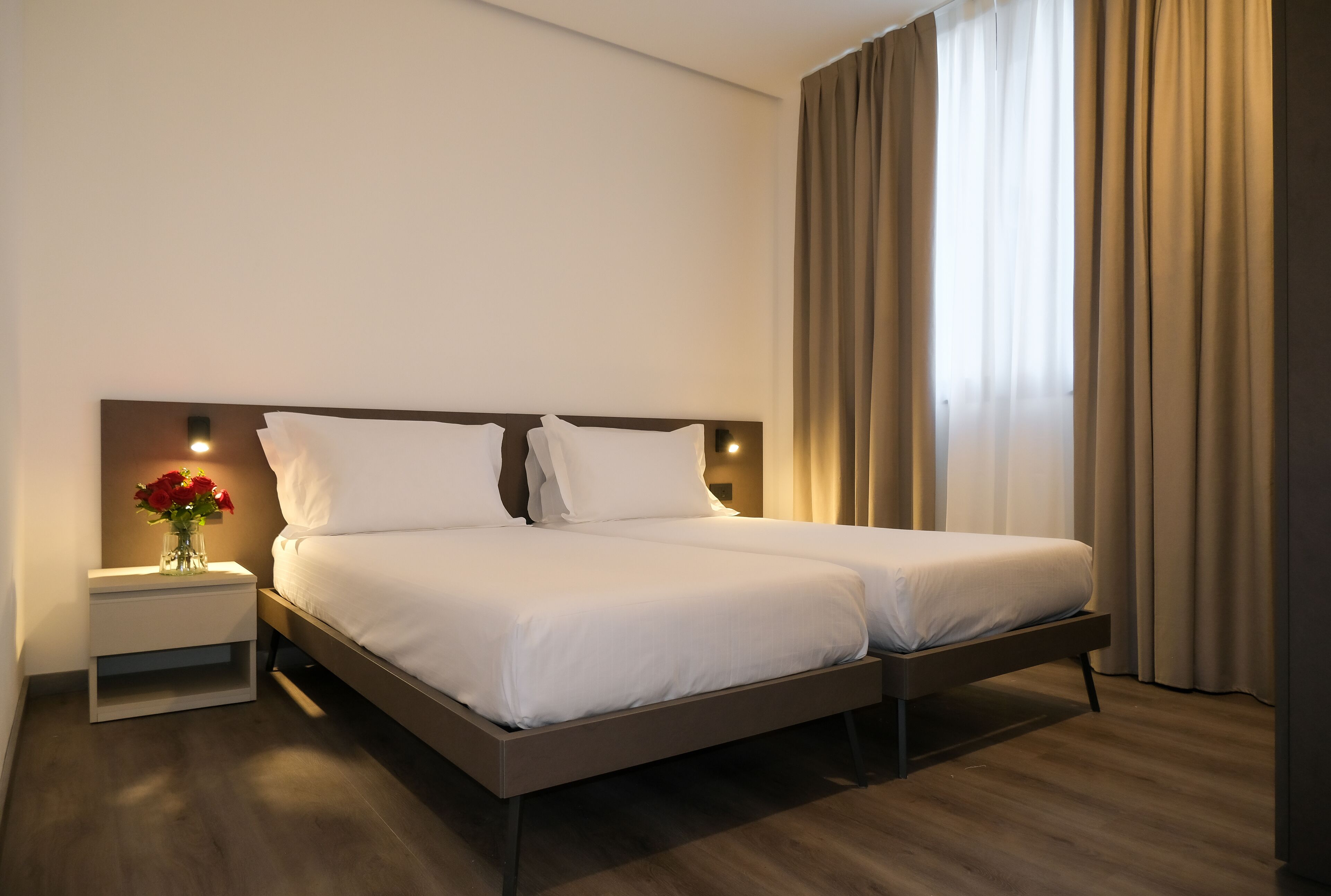 Ein Doppelbett mit weißer Bettwäsche, ein Nachttisch mit einer Vase roter Blumen und ein großes Fenster mit Vorhängen.