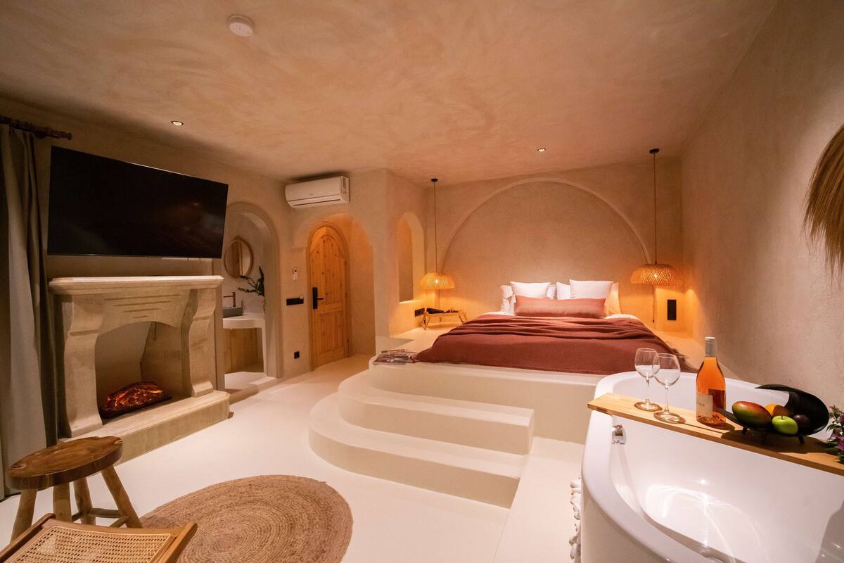 A bedroom with a fireplace, a large bed, a bathtub, and a tray with glasses and fruit.
