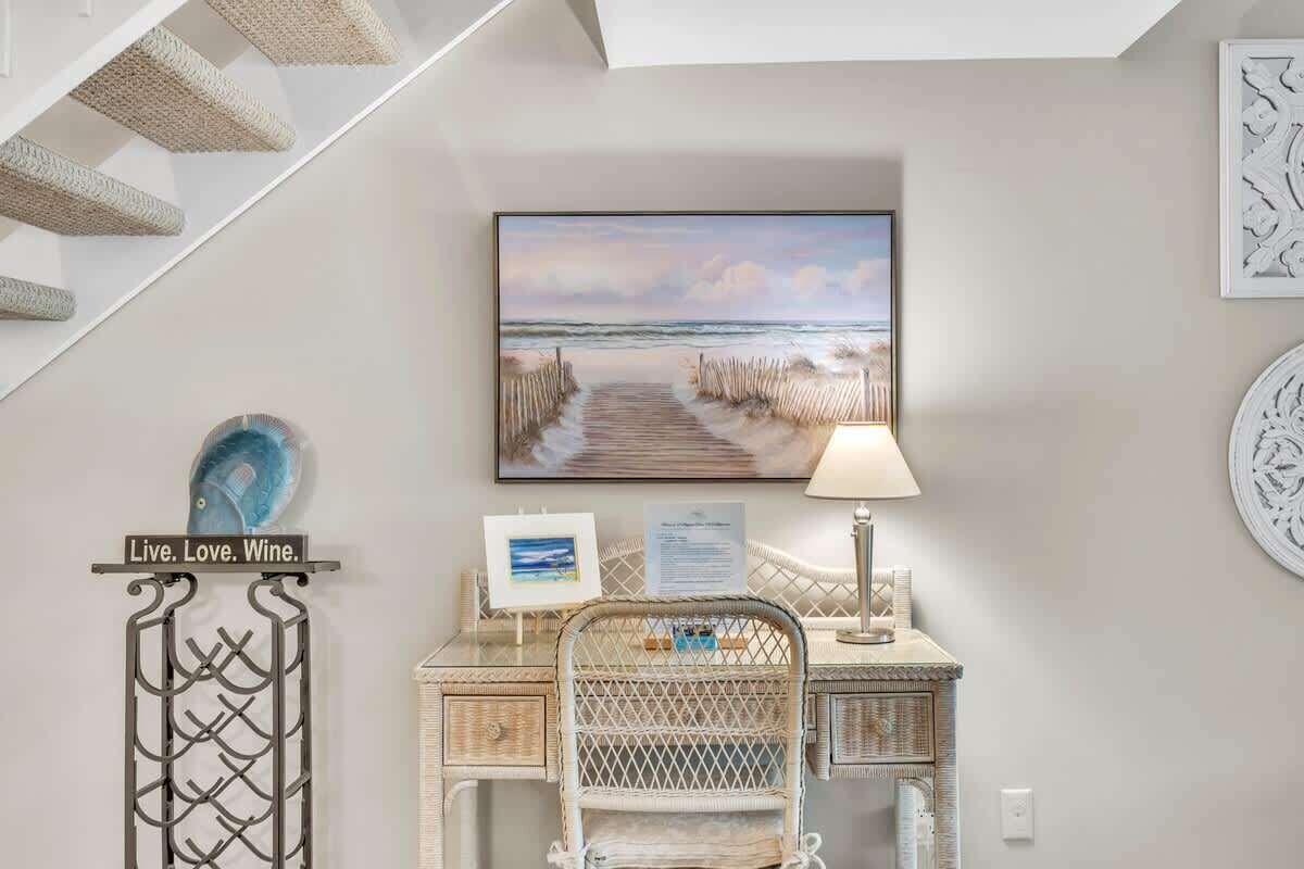 A room with a desk, a chair, a framed picture of a beach, a lamp, and a sign that says 'Live. Love. Wine.'