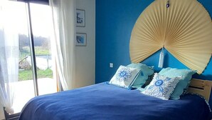 A bedroom with a bed, a blue wall, a large fan, and a view of the outdoors.